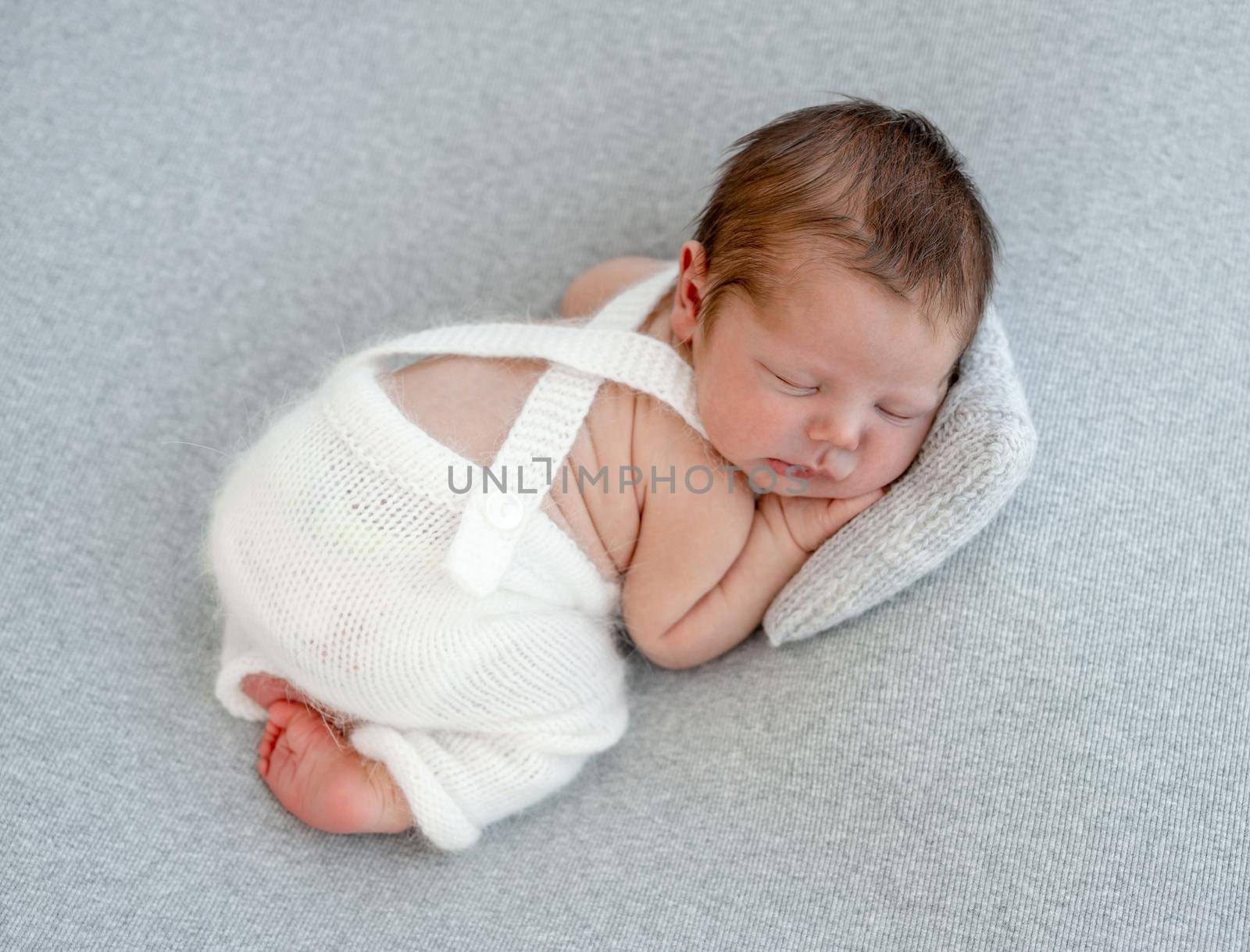 Newborn baby boy sleeping wearing knitted white costume and holding his hands under cheeks. Cute infant kid studio portrait with pillow