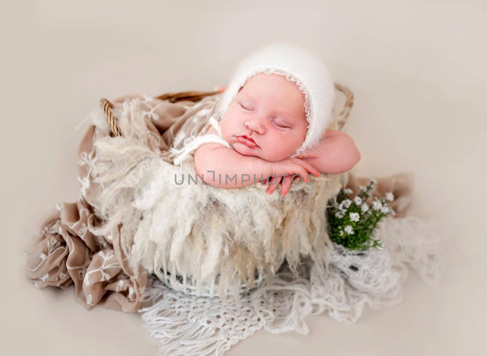 Newborn baby girl wearing knitted white hat sleeping in the basket and holding her hands under cheek. Little cute infant child napping. Adorable kid during studio photoshoot on beige background