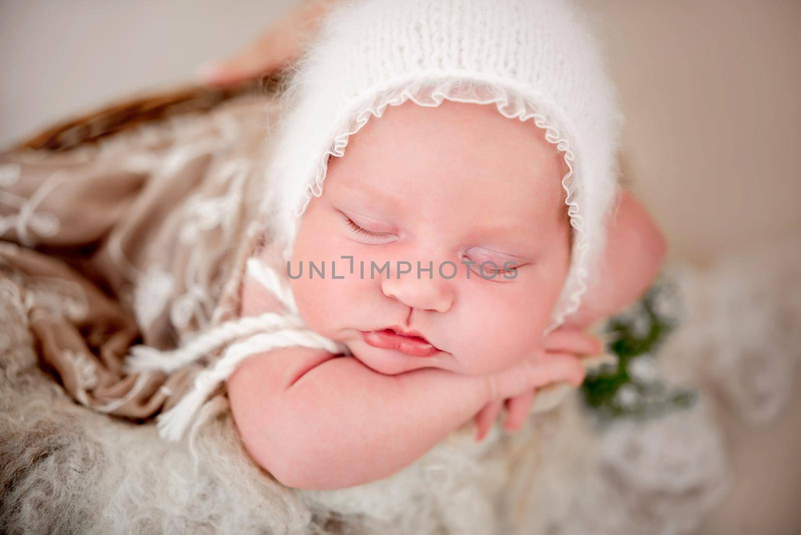 Closeup portrait of newborn baby girl wearing knitted white hat sleeping in the basket and holding her hands under cheek. Little cute infant child napping