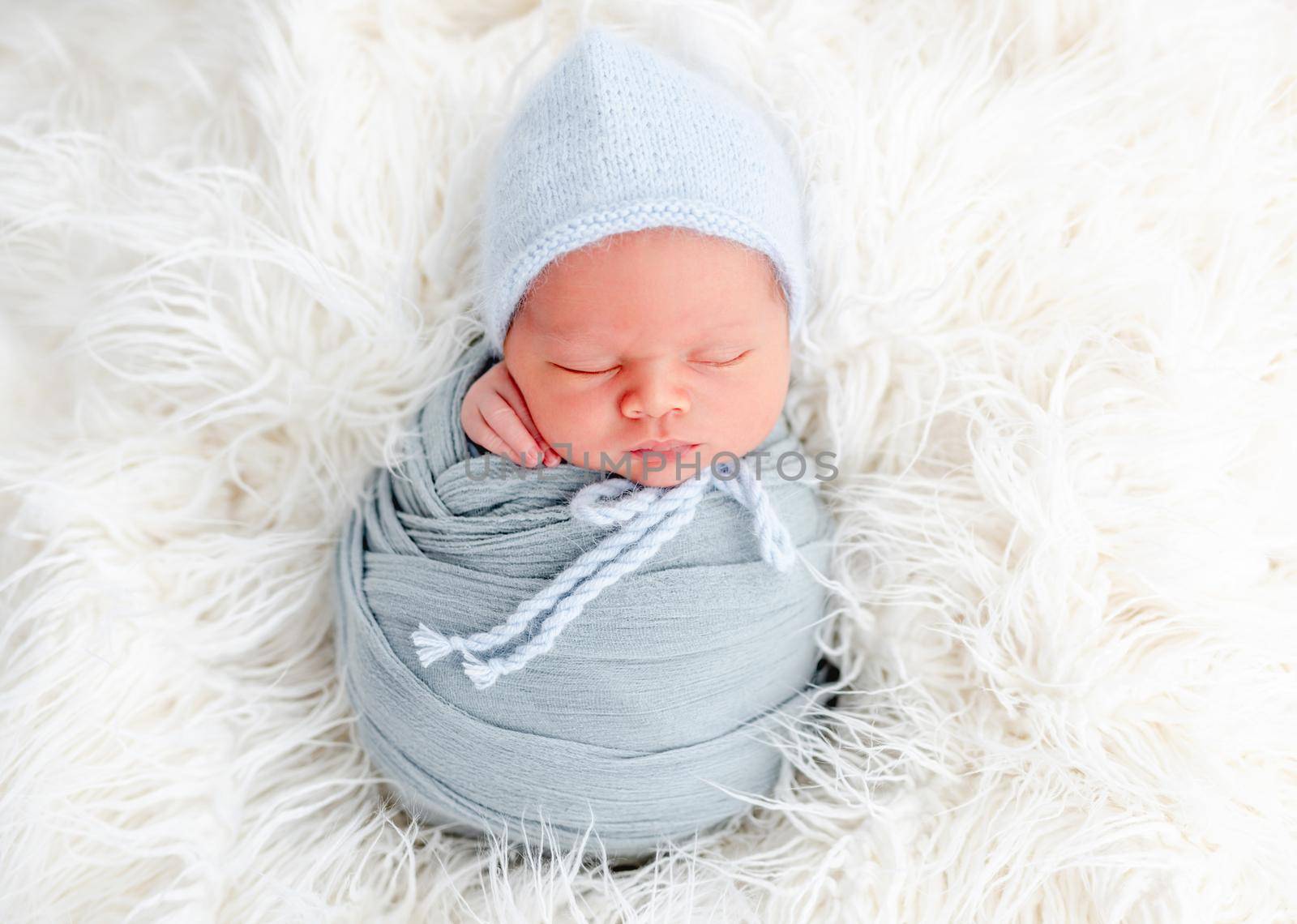 Portrait of newborn baby boy swaddled in light blue fabric and wearing knitted hat sweet sleeping on white fur. Adorable infant child napping