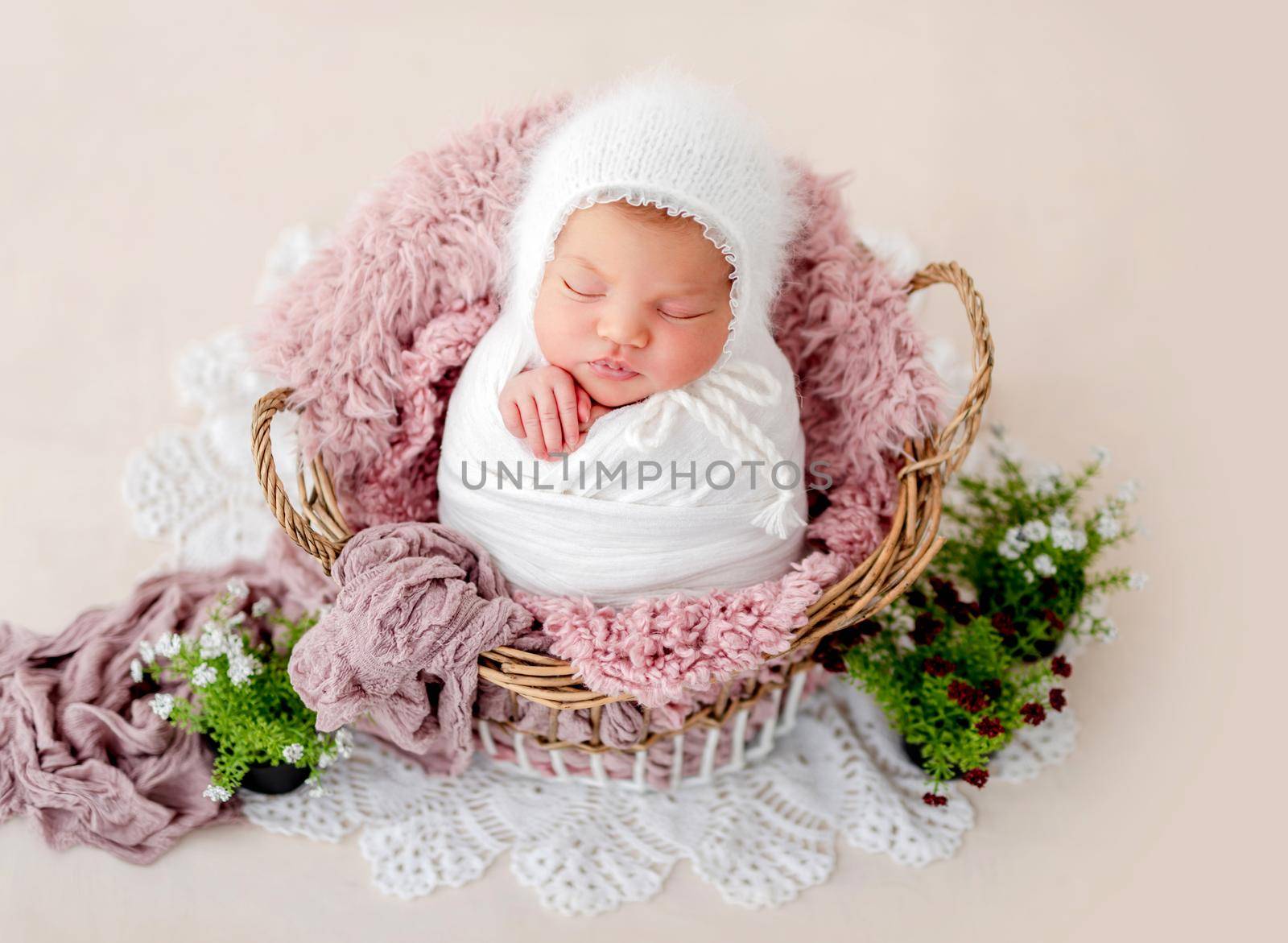 Swaddled beautiful newborn baby girl sleeping in the basket with fur on light pink background. Cute female infant child photoshoot in studio. Adorable kid napping