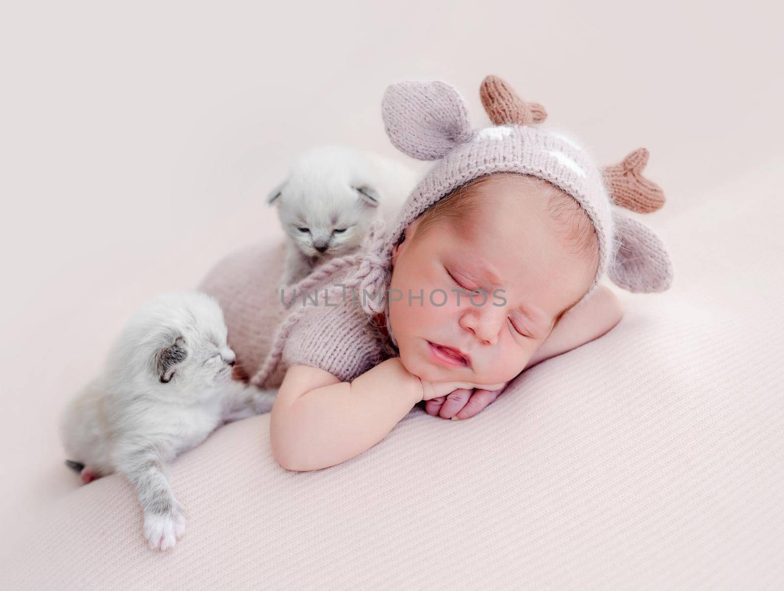 Adorable newborn baby boy sleeping on his tummy and two little fluffy kittens sitting close to him. Cute infant kid wearing knitted costume and hat napping with cats during studio photoshoot