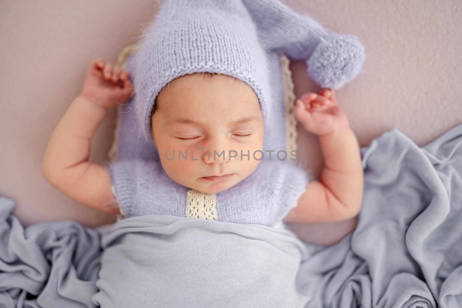 Beautiful little newborn baby girl wearing knitted costume and hat sleeping with hands up under blanket in studio. Cute infant child napping