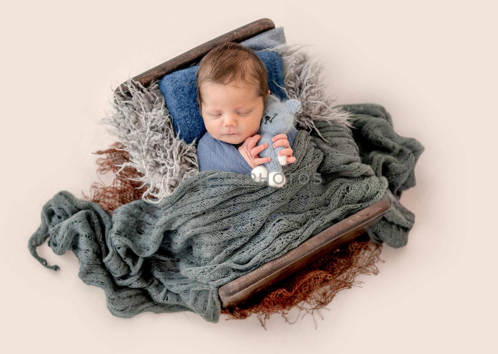 Newborn baby boy sleeping in tiny bed in knitted blanket and hugging toy. Infant kid studio portrait with decoration