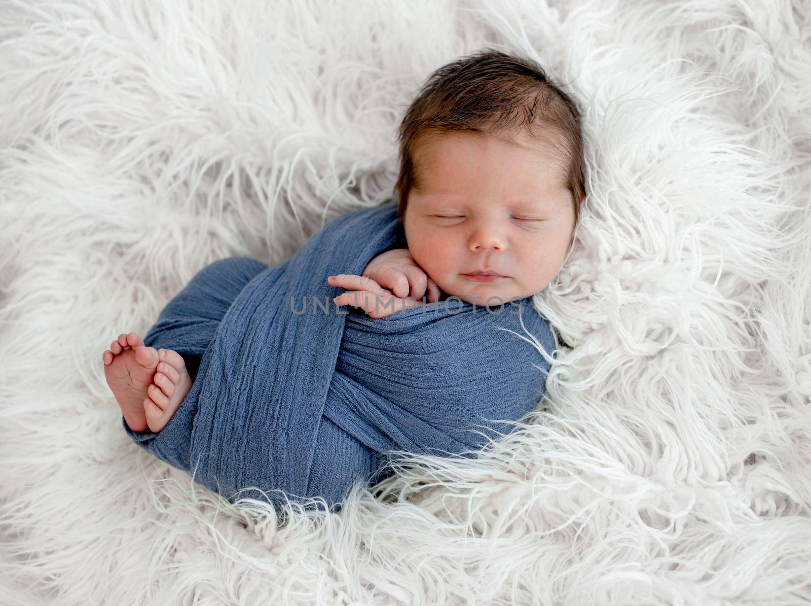 Newborn baby boy sleeping swaddled in fabric on white fur. Infant kid studio portrait with decoration