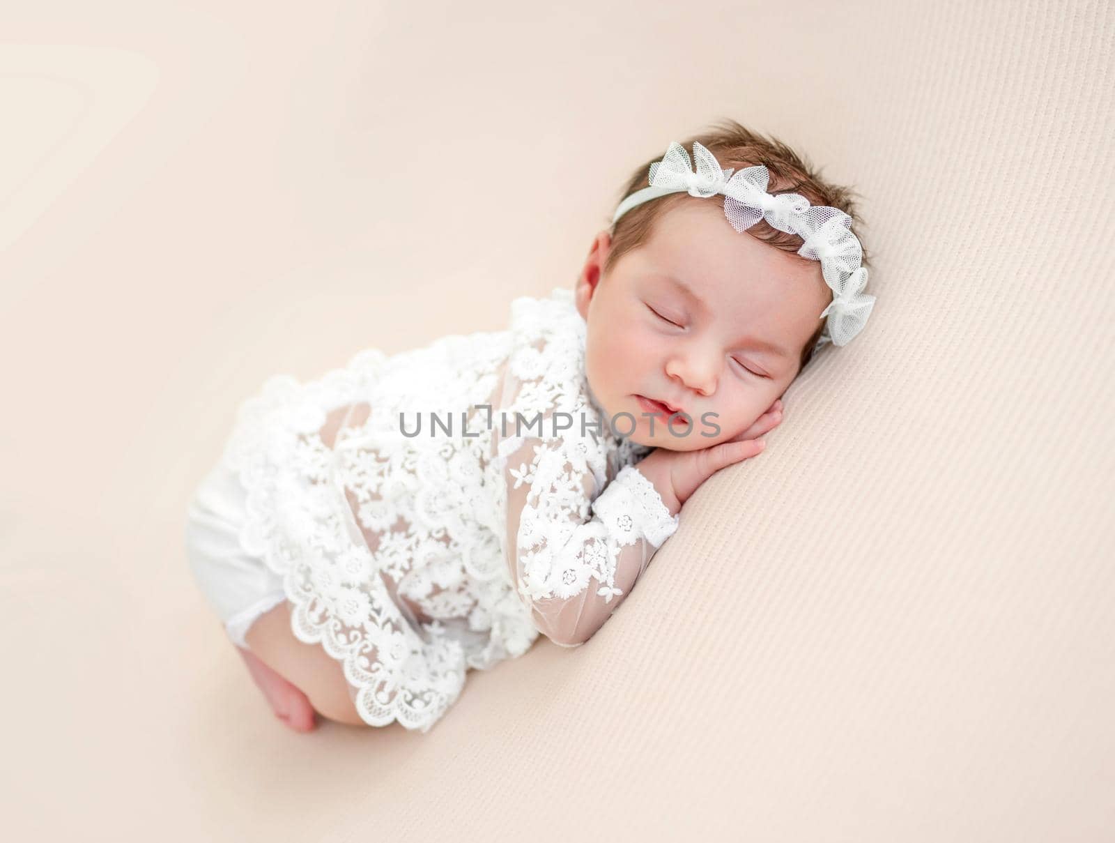Adorable newborn baby girl wearing beautiful white costume and wreath lying on her tummy and sleeping in studio. Cute infant child napping holding hands under cheeks