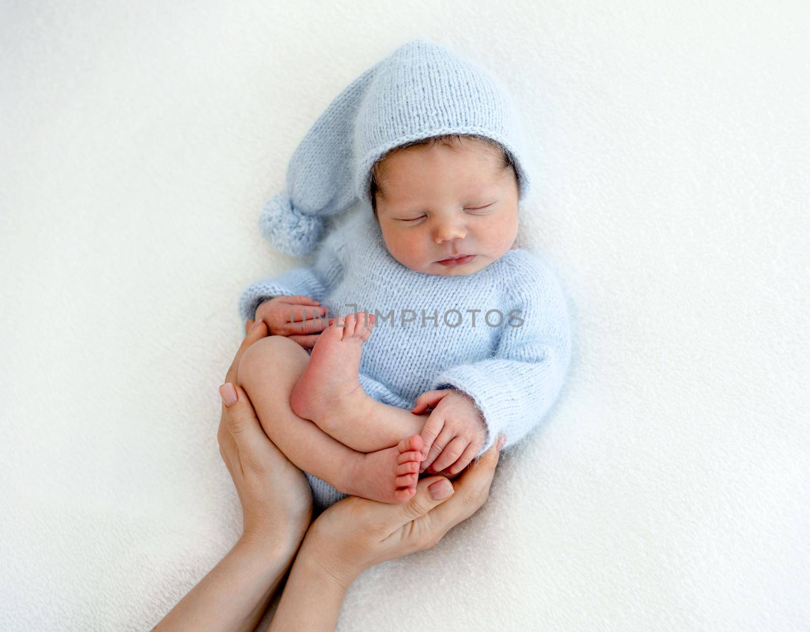 Sweet newborn baby boy sleeping wearing knitted costume and his mother hands care about him. Napping infant kid studio portrait with parent
