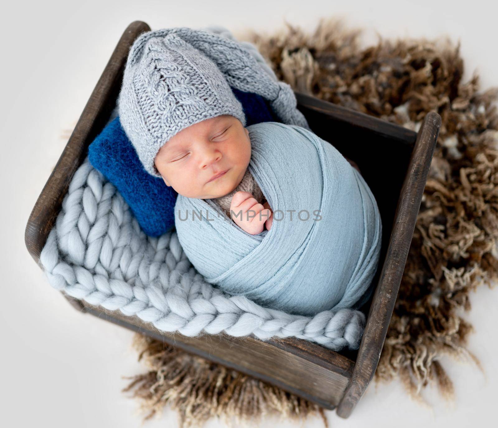 Adorable newborn baby boy swaddled in fabric and wearing cute knitted hat sleeping in wooden stylized bed in studio. Infant child napping indoors