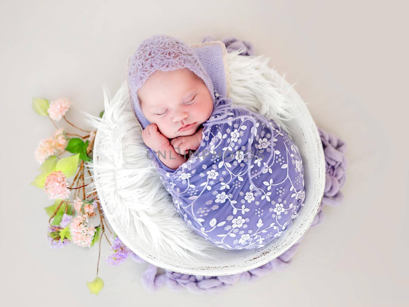 Newborn baby girl swaddled in beautiful fabric and wearing knitted hat sleeping in white basin. Little cute infant child napping lying on white fur. Adorable kid during studio photoshoot