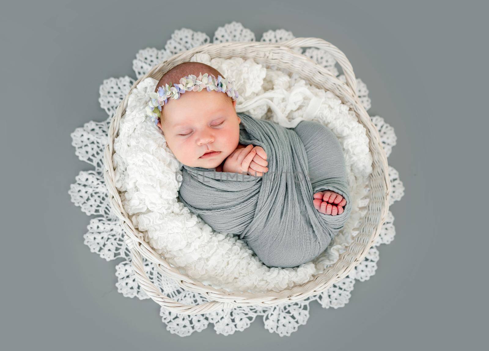 Adorable newborn baby girl wearing wreath lying on her back in basket and sleeping. Sweet swaddled in fabric infant child napping during studio photoshoot with decoration