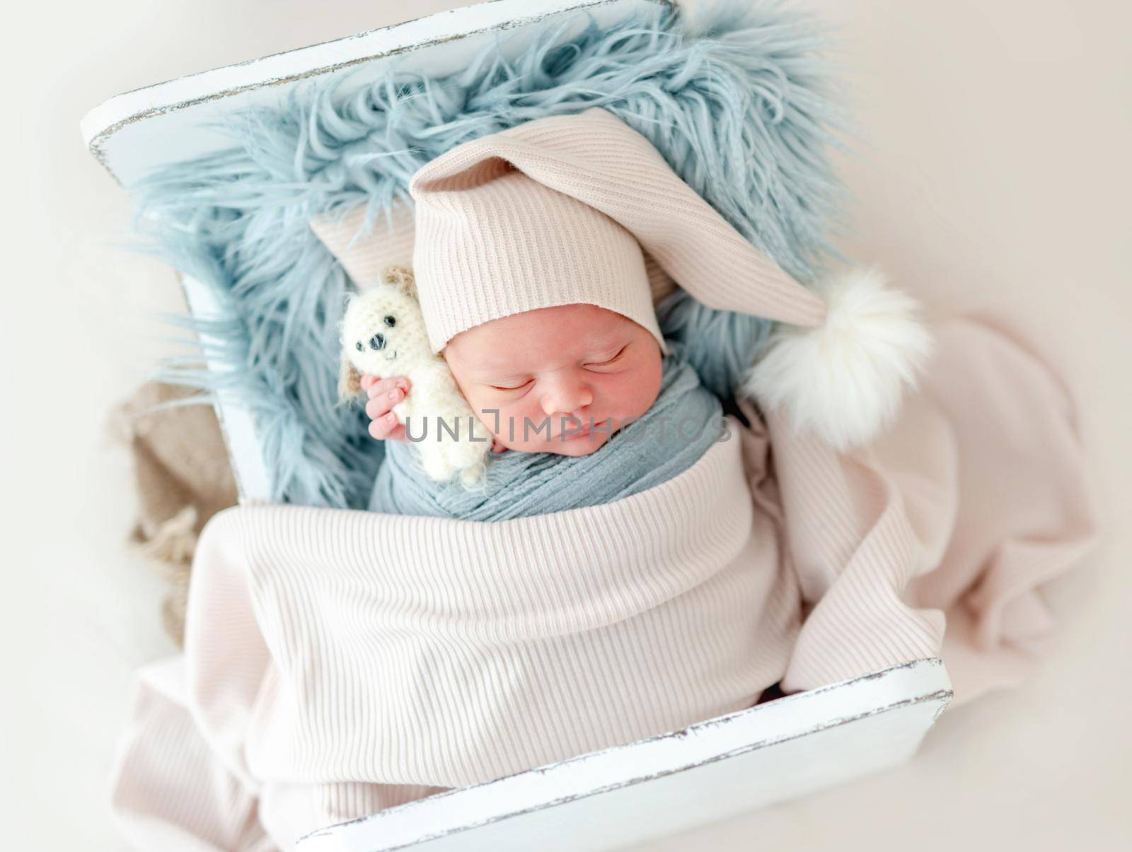 Portrait of newborn baby boy wearing knitted hat sleeping on white small designed bed under blanket and holding toy. Adorable infant child napping during studio photoshoot