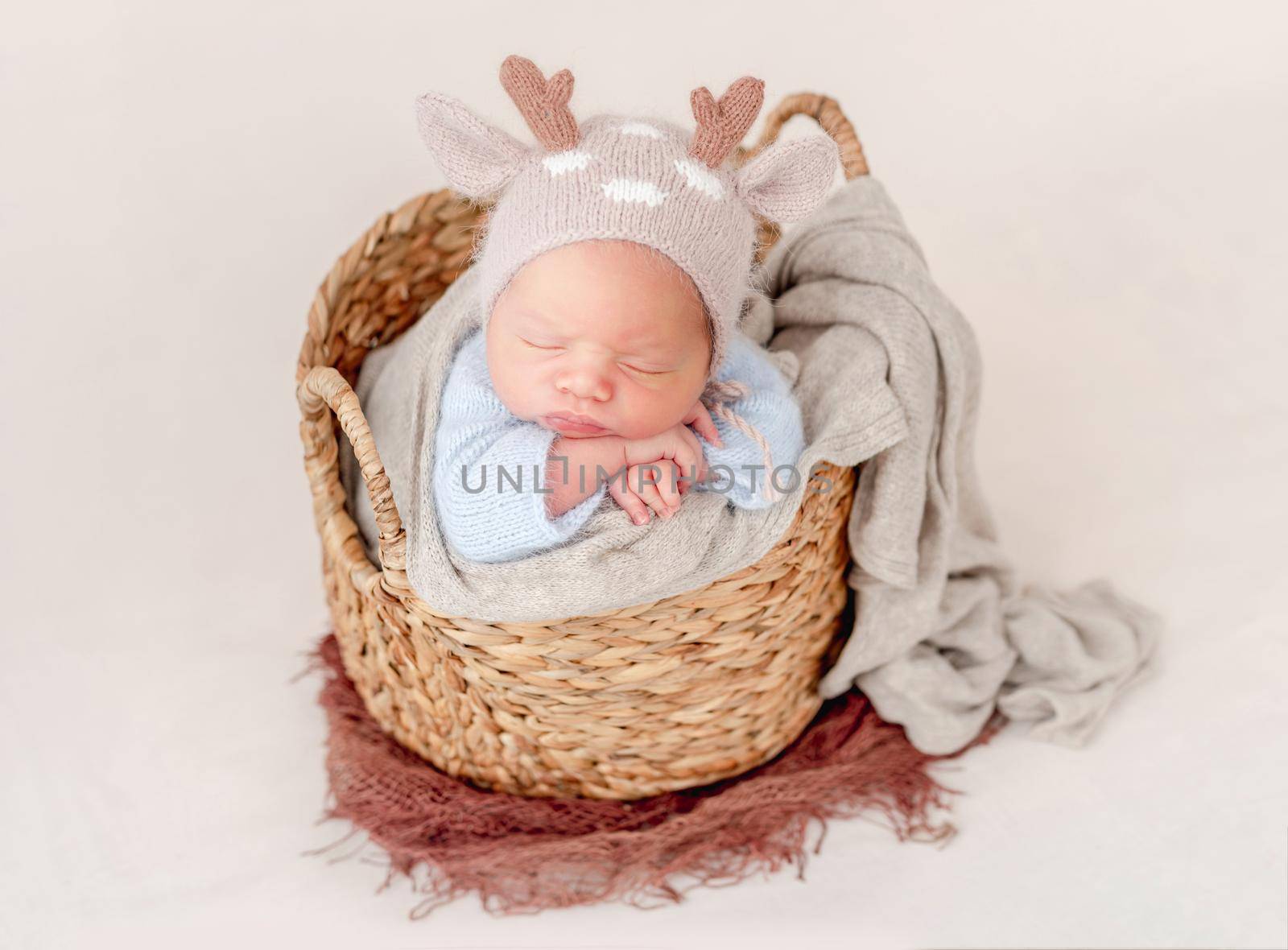 Portrait of newborn baby boy wearing knitted hat with deer horns sleeping on his tummy in basket and holding tiny hands under his cheeks. Adorable infant child napping in studio