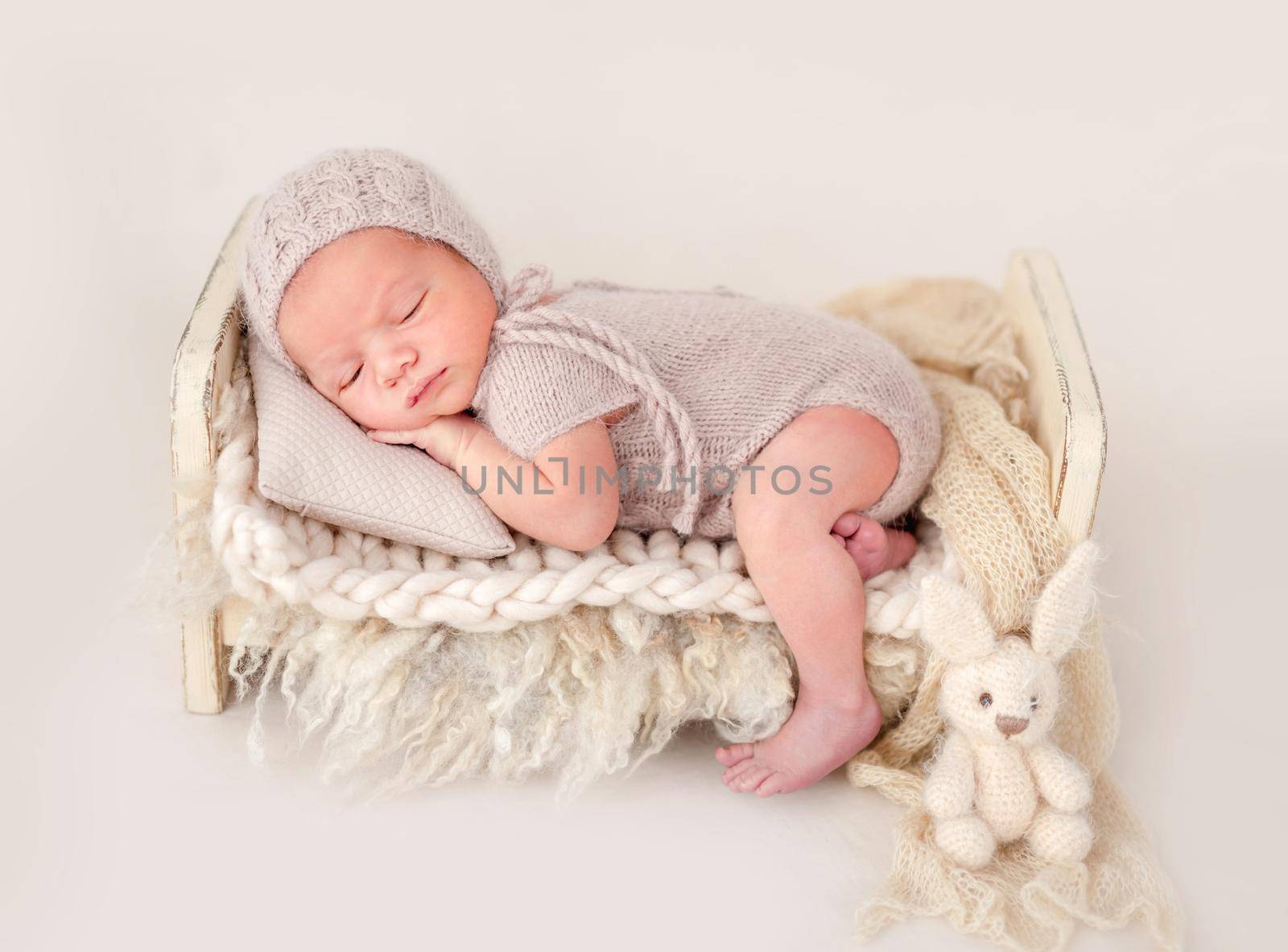 Newborn baby boy wearing knitted beige costume and hat sleeping on his tummy in small designed bed and holding tiny hands under his cheeks. Adorable infant child napping