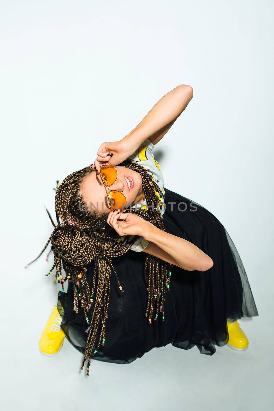 Portrait of a young stylish woman with braided hair dressed in white t-shirt, black skirt, yellow boots and sunglasses posing on the white background.