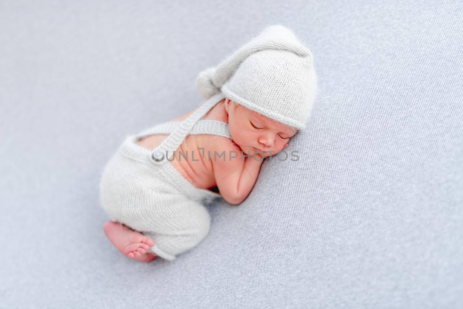 Newborn baby boy wearing knitted pants and hat sleeping on his tummy and holding tiny hands under his cheeks. Adorable infant child napping in studio with light blue colors