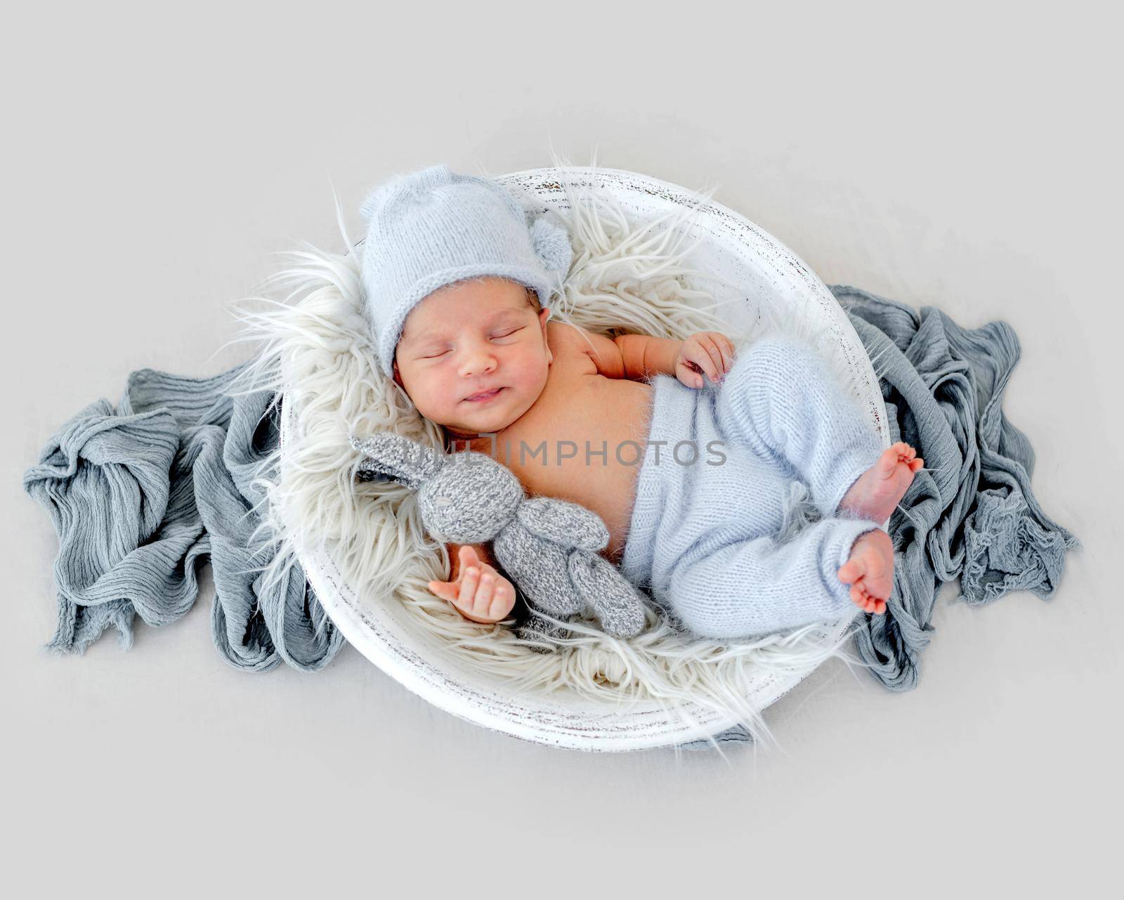 Adorable newborn baby boy wearing cute knitted hat and pants sleeping in wooden basin on fur and holding handmade bunny toy. Infant child napping