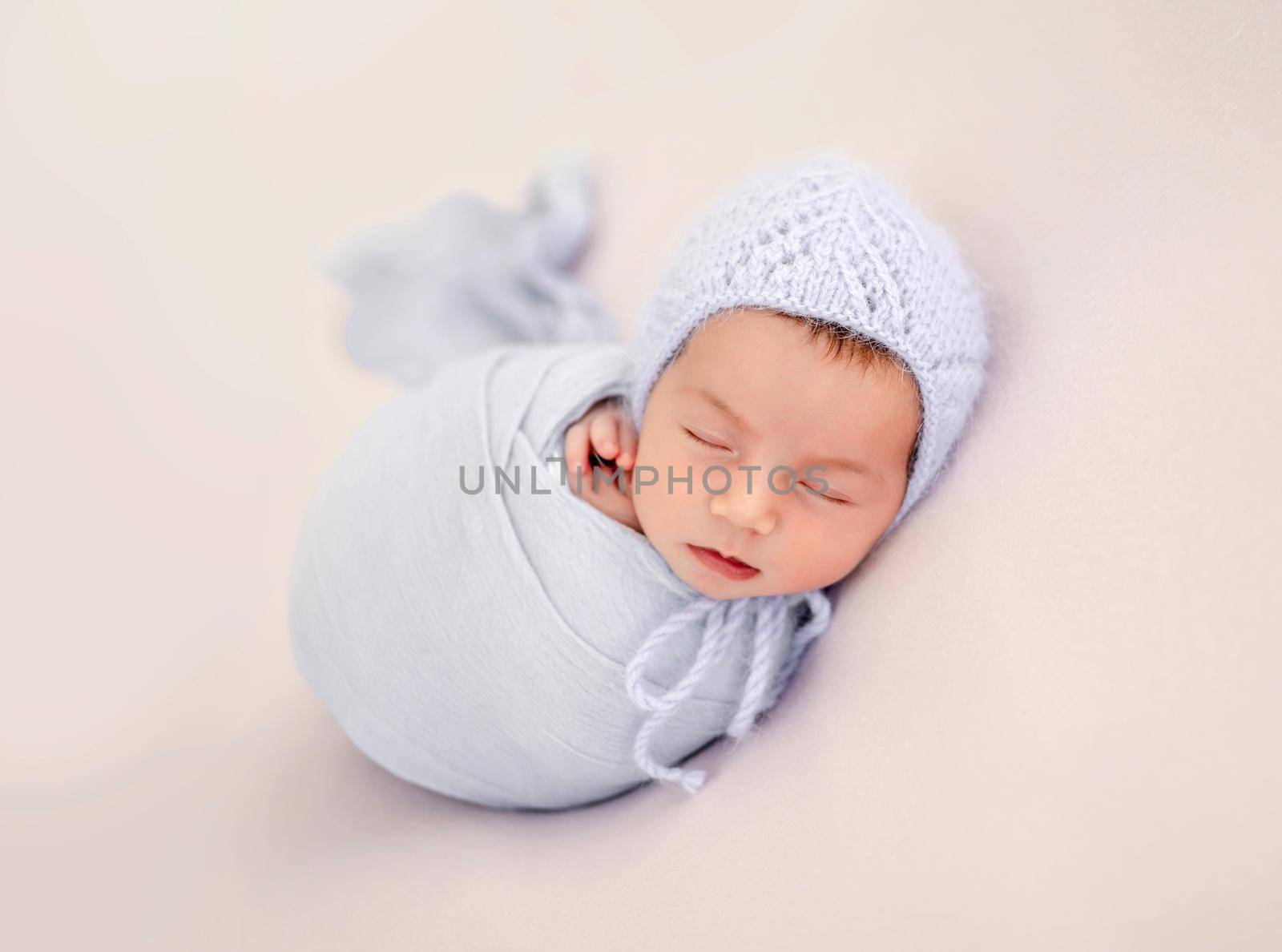 Little beautiful newborn baby girl swaadled in fabric and wearing knitted hat sleeping during studio photoshoot. Cute infant child kid napping resting