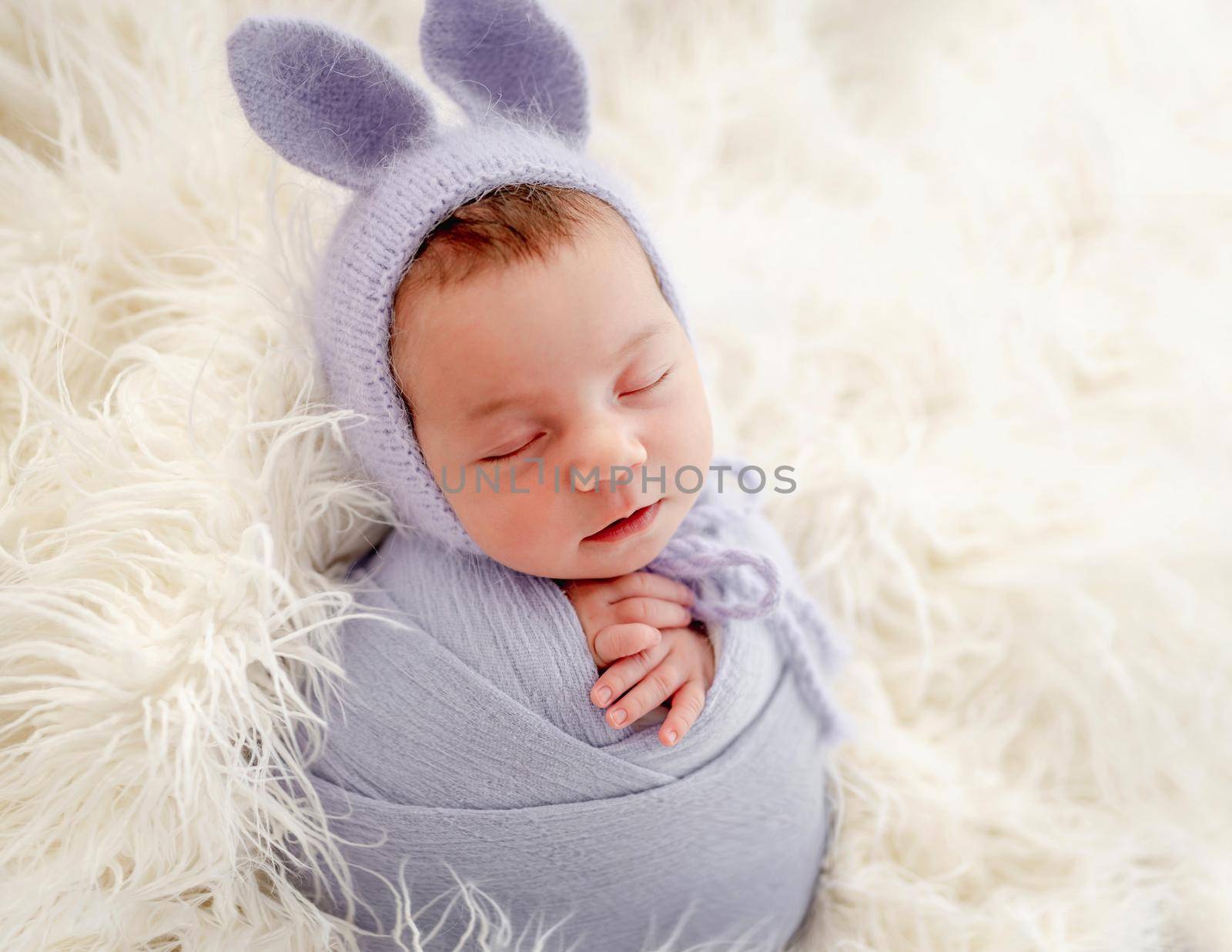 Little beautiful newborn baby girl swaadled in fabric and wearing hat with bunny ears sleeping on fur during studio photoshoot. Cute infant child kid napping