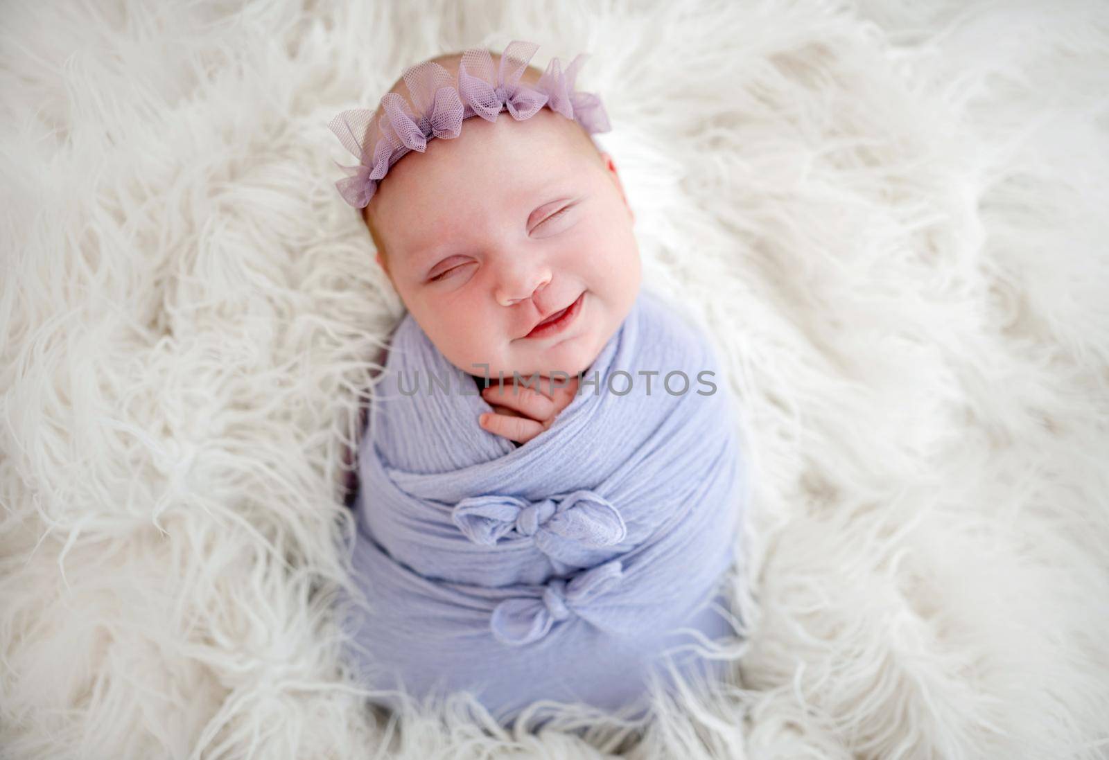 Newborn baby girl swaddled in light blue fabric sleeping and smiling. Little cute infant child napping lying on white fur. Adorable kid during studio photoshoot