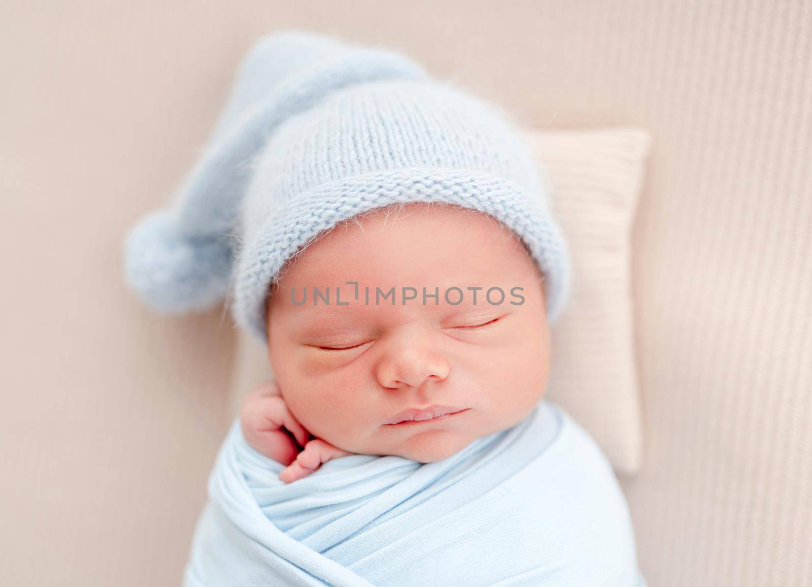 Portrait of newborn baby boy swaddled in light blue fabric and wearing knitted hat sleeping on white background and holding tiny hands under his cheeks. Adorable infant child napping