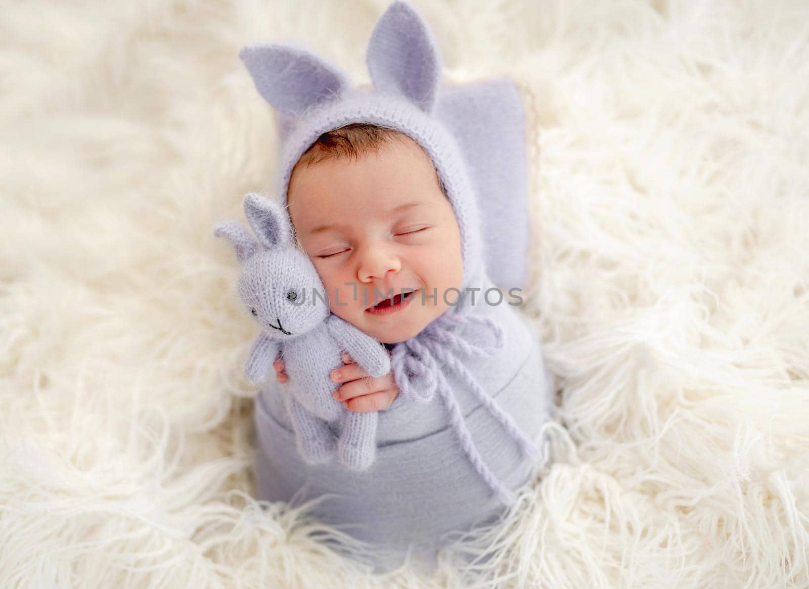 Little beautiful newborn baby girl swaadled in fabric and wearing hat with bunny ears sleeping on fur and smiling during studio photoshoot. Cute infant child kid napping holdint toy in tiny hands