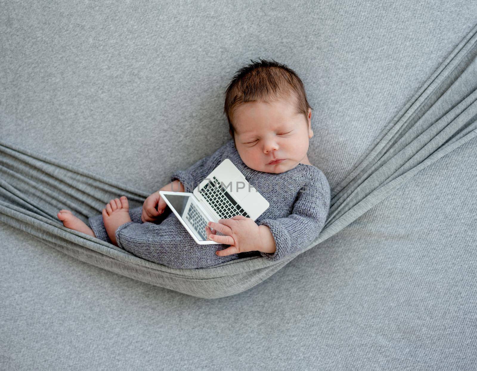 Newborn baby boy wearing in knitted costume sleeping in hammock and holding toy laptop. Infant kid studio portrait with decoration