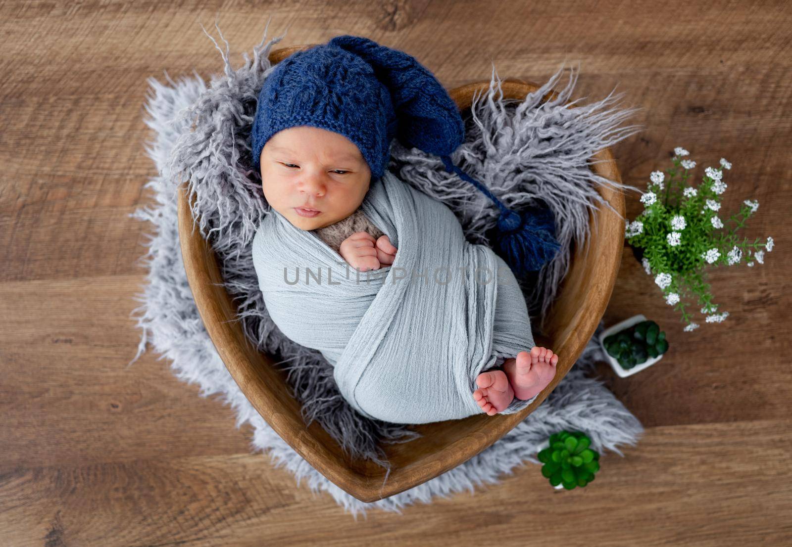 Adorable newborn baby boy swaddled in fabric and wearing cute knitted hat falling asleep during studio photoshoot in wooden heart shape bed. Infant child napping