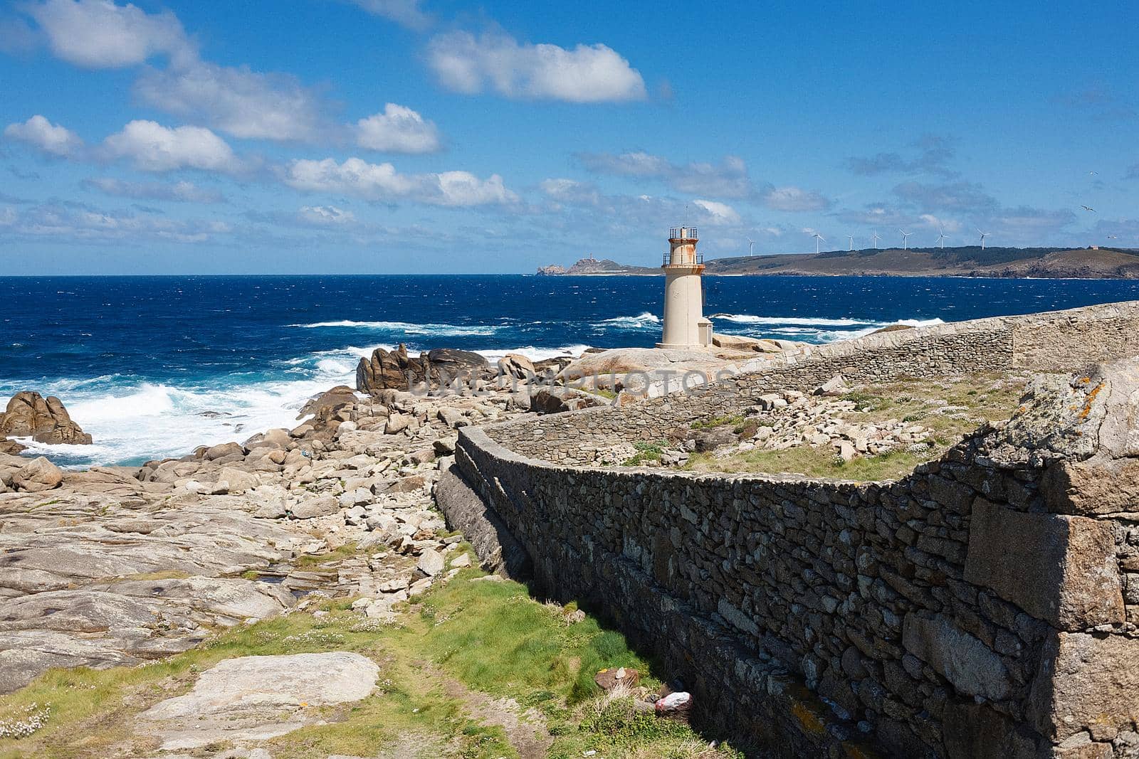 View on a lighthouse and ancient wall made of stones near the ocean with blue skies and white clouds. The scene from Camino de Santiago, tourist path in Spain.