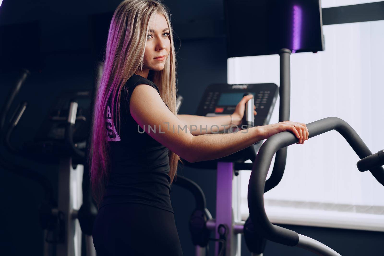 Attractive young blonde woman exercising on cardio training apparatus in dark gym
