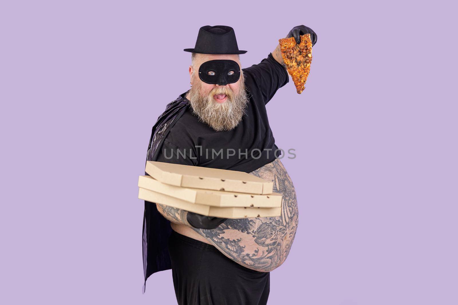 Funny plump man in hero suit with large bare abdomen holds boxes stack and slice of tasty pizza standing on purple background in studio