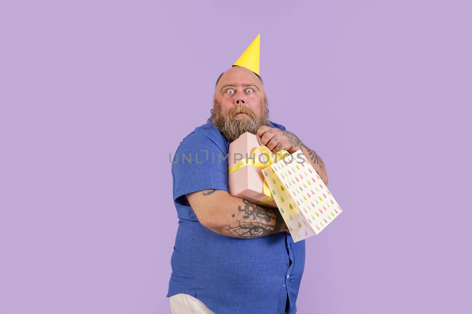 Funny scared mature man with overweight in yellow party hat holds presents standing on purple background in studio