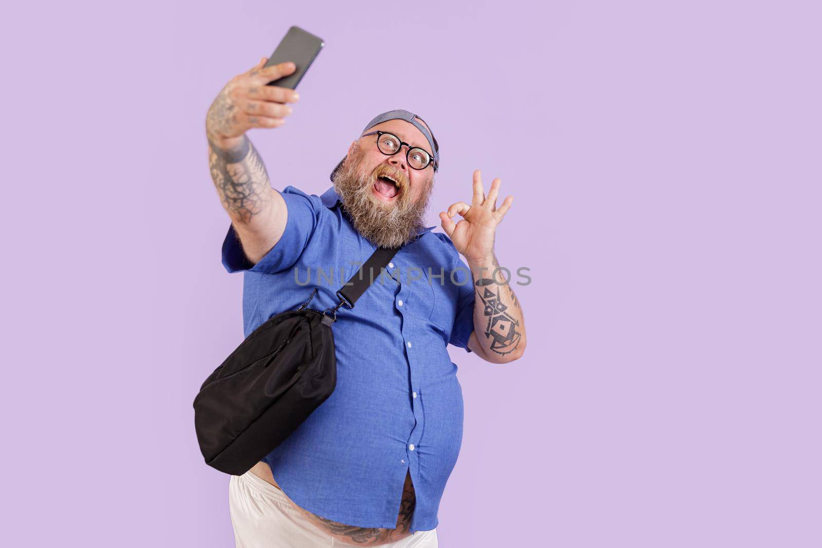 Emotional plump hipster man with crossbody bag shows Ok gesture taking selfie with modern mobile phone on purple background in studio