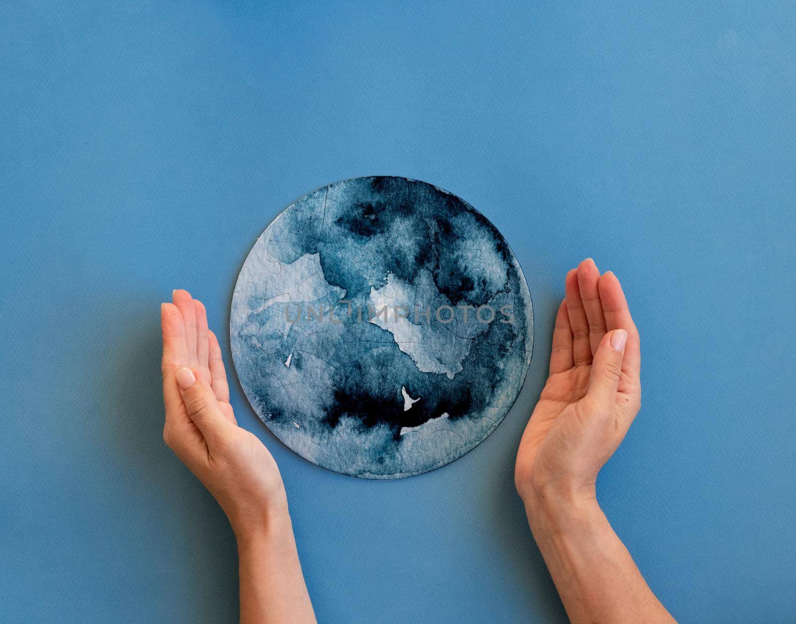 Hands holding unfinished jigsaw puzzle in shape of globe on blue background. Protecting the planet concept