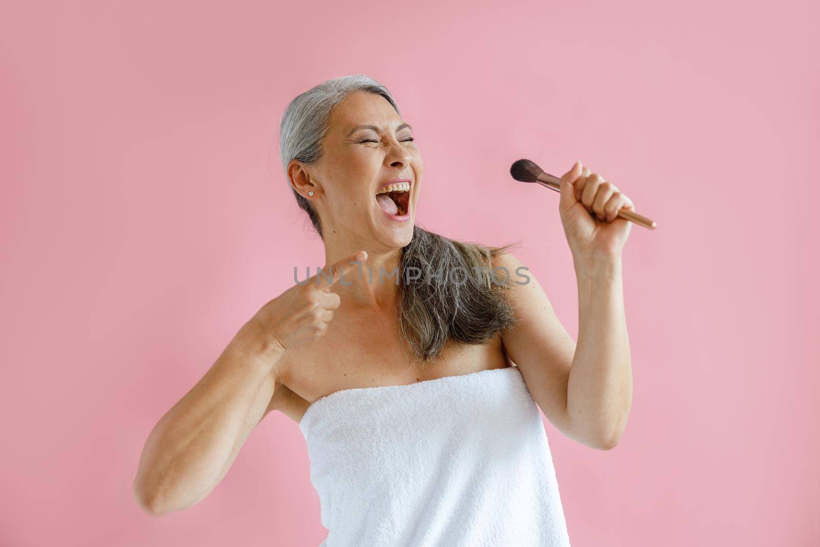 Excited middle aged Asian model wrapped with terry towel sings song holding cosmetic brush on pink background in studio. Mature beauty lifestyle