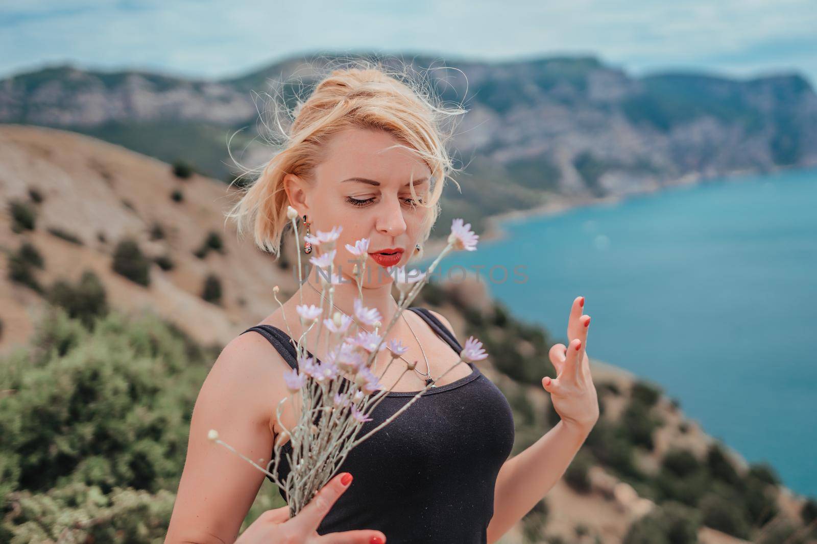 blonde girl with scarlet lipstick and red manicure in a black T-shirt with a bouquet of wildflowers on a background of sea and mountains. she is happy and enjoying wonderful breathtaking landscape.