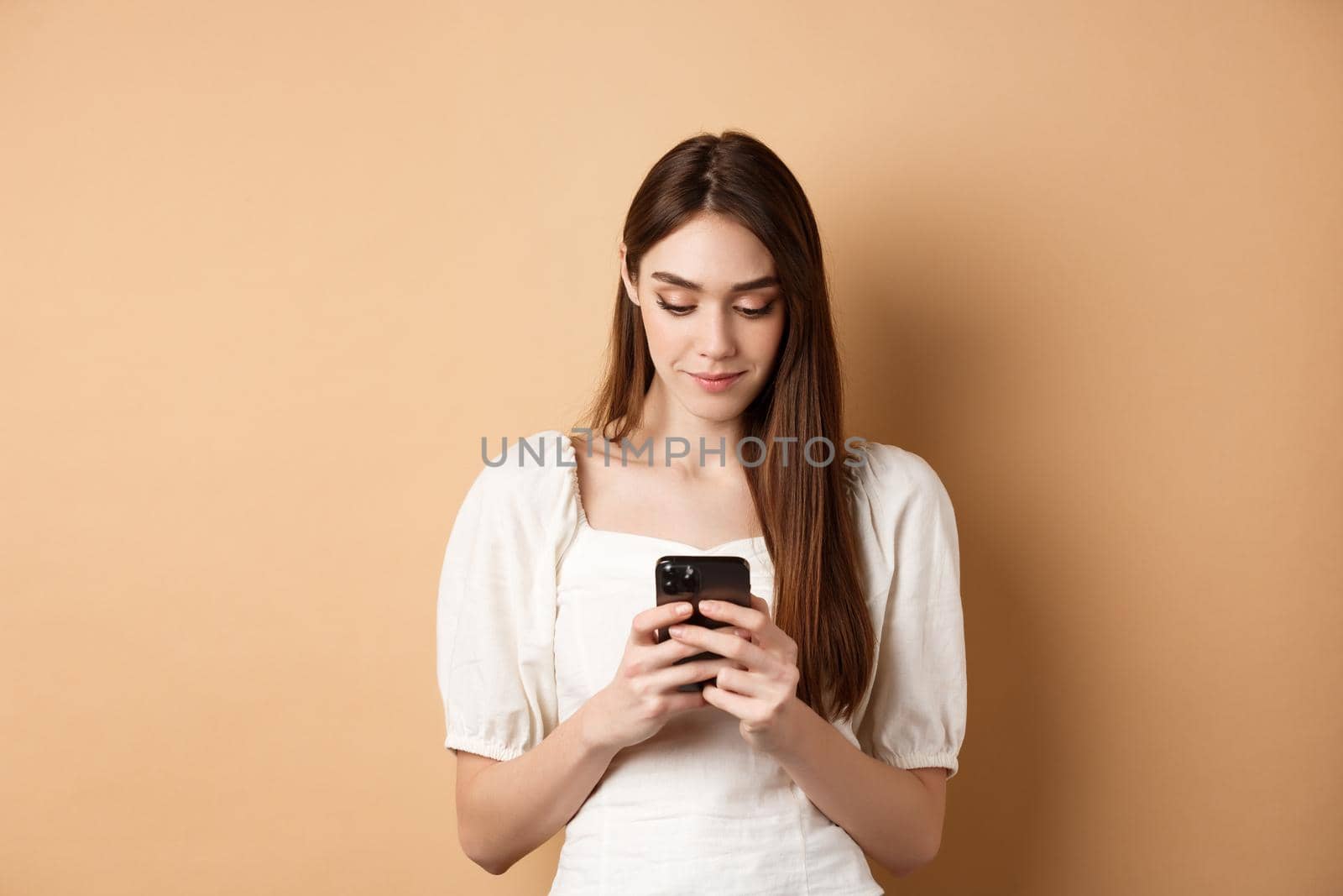 Pretty woman texting on smartphone cell phone, reading screen and smiling, standing on beige background.