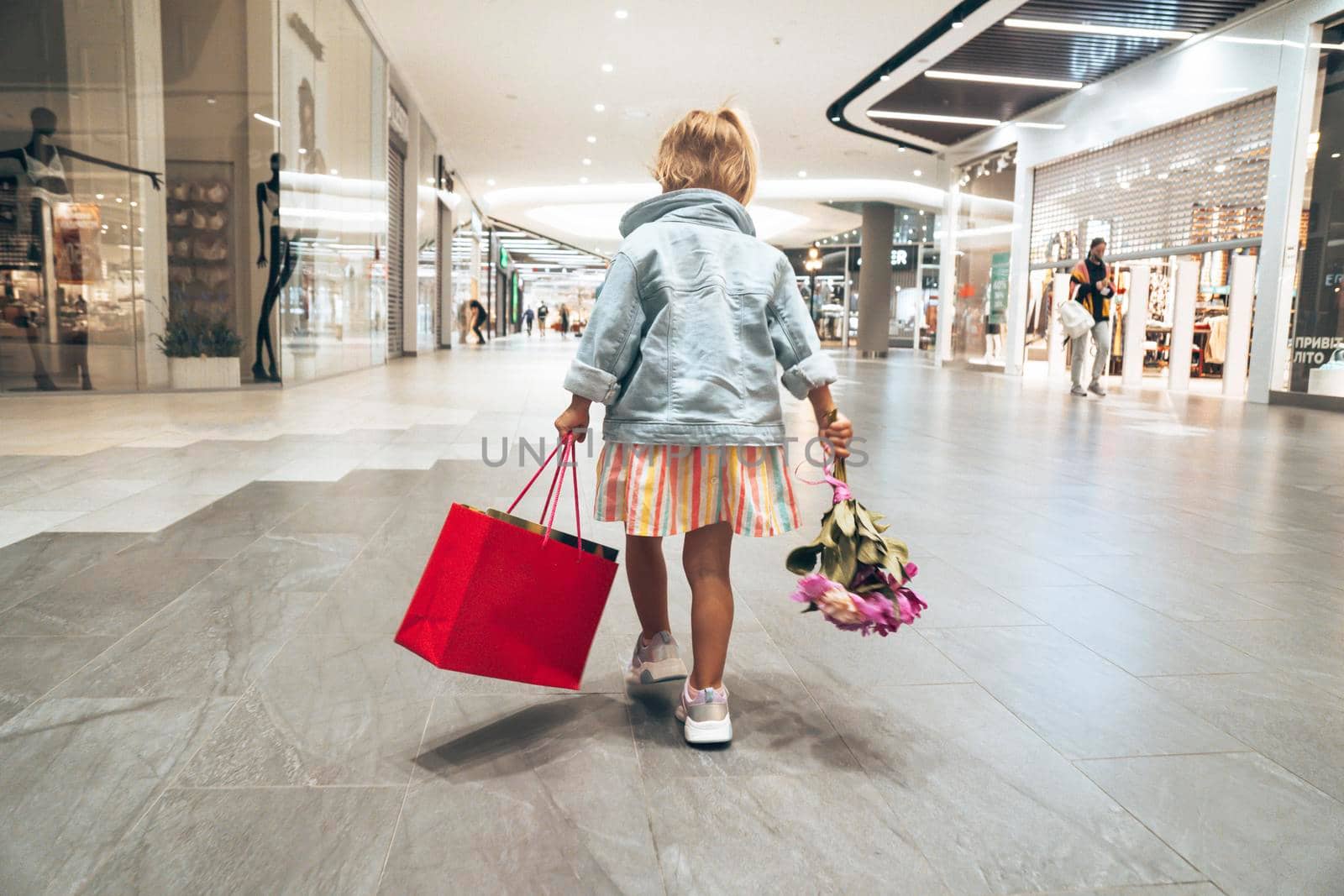 Small cute fashion child is carrying shopping bag and flowers inside the Shopping Mall after black Friday sale. View from behind. Toddler is happy and feels she is a shopoholic.