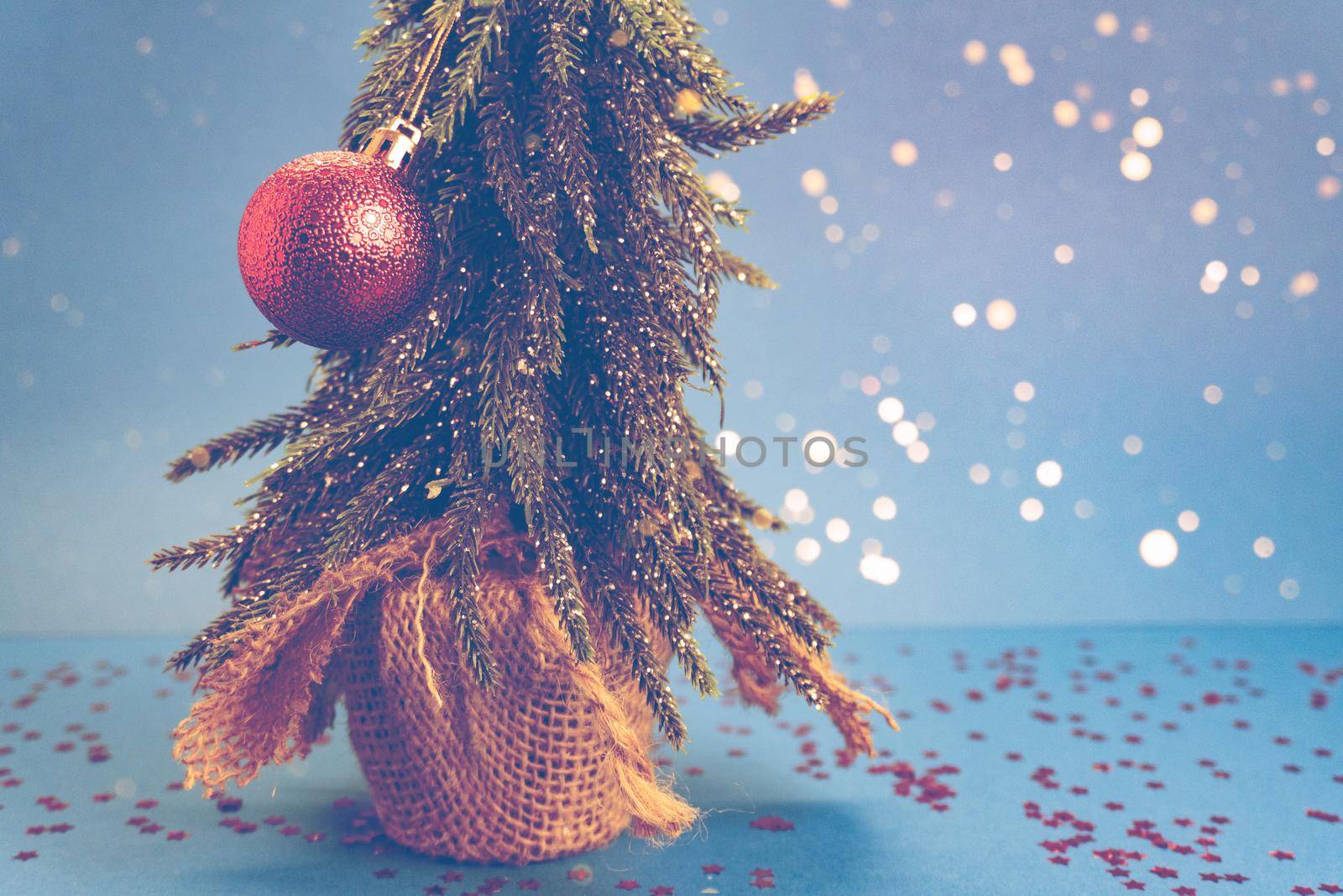 Christmas tree with red fragile toy is on blue table with red stars and blue background behind the tree. New year and christmas, romantic and family concept, bokeh. Copyspace for the text. Toned