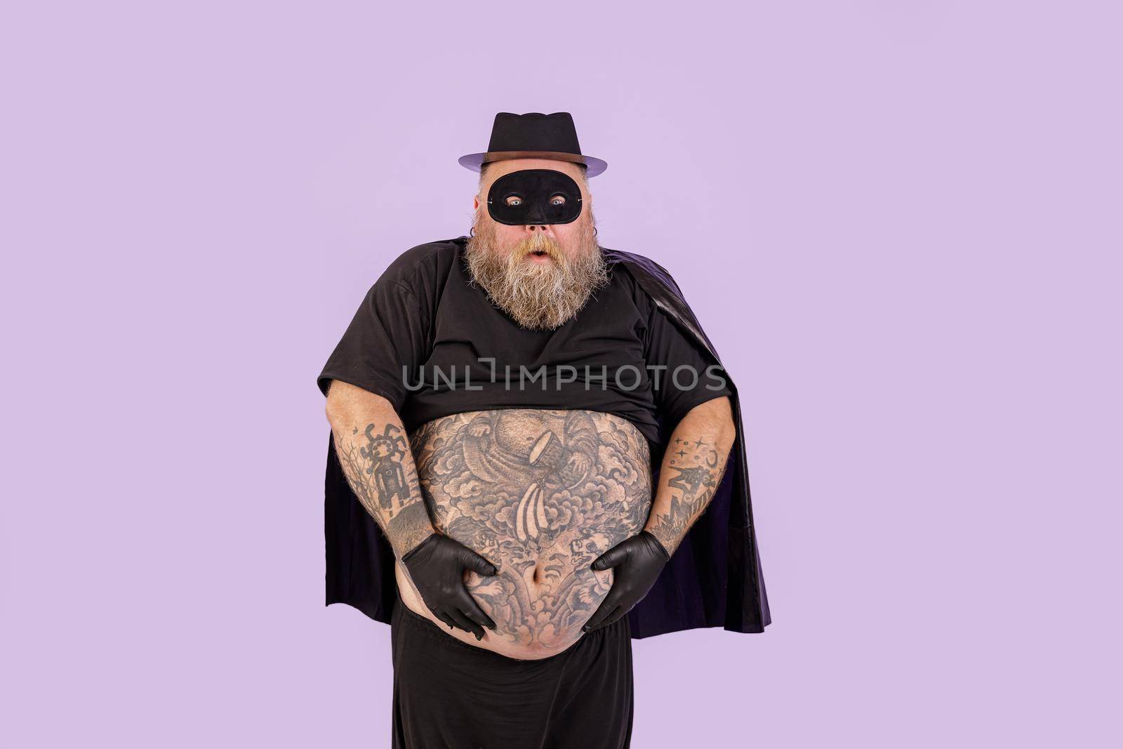Doubting bearded man with overweight wearing carnival suit with cape and mask holds huge belly covered with tattoos standing on purple background in studio