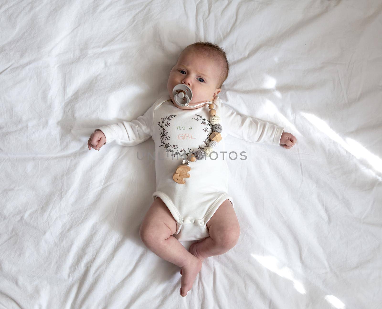 Portrait of a newborn baby girl who lies on a bed with a nipple soother. Three-month-old girl in white suit lies on a veil view from above. High quality photo