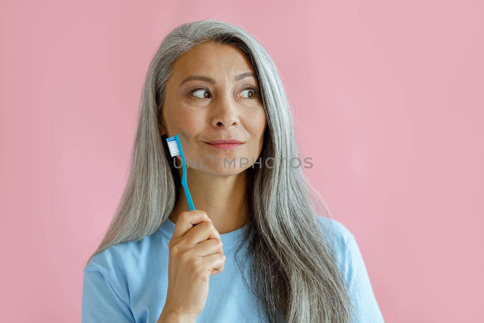 Positive thoughtful middle aged Asian woman holds toothbrush in studio by Yaroslav_astakhov