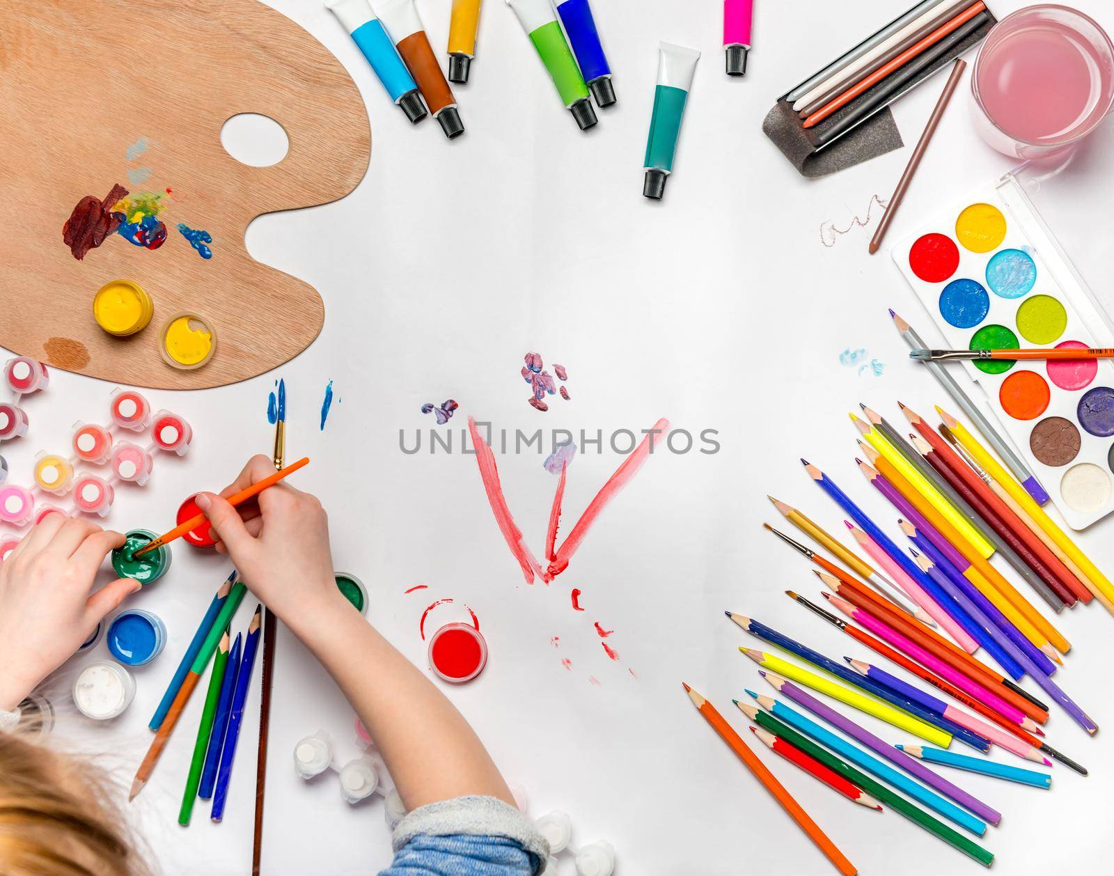 Top view of girl's hand with pencil over white blank paper and colorful pencils and watercolors on white wooden table background. Copy space