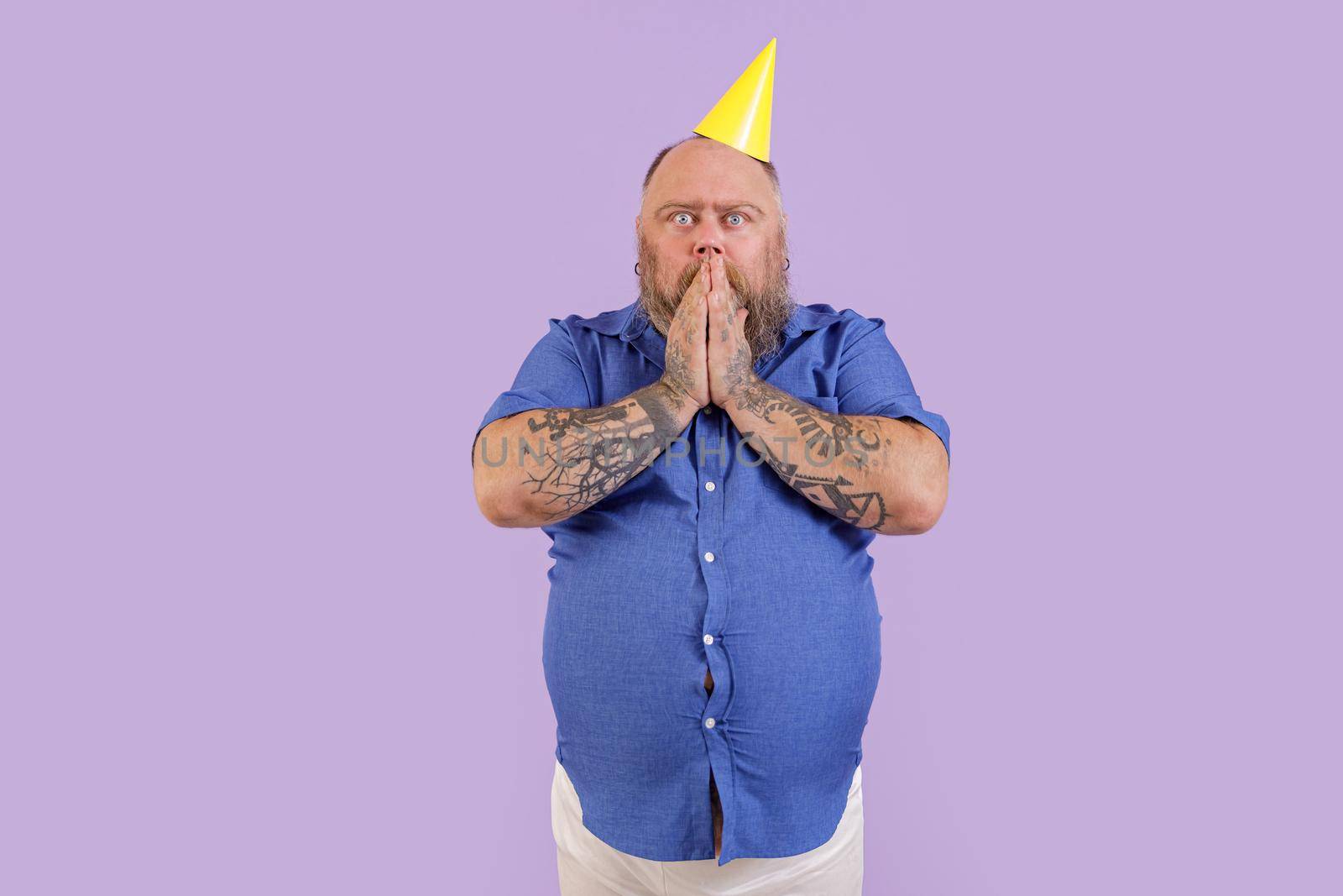Expressive surprised bearded man with overweight in tight shirt holds hands over mouth standing on purple background in studio