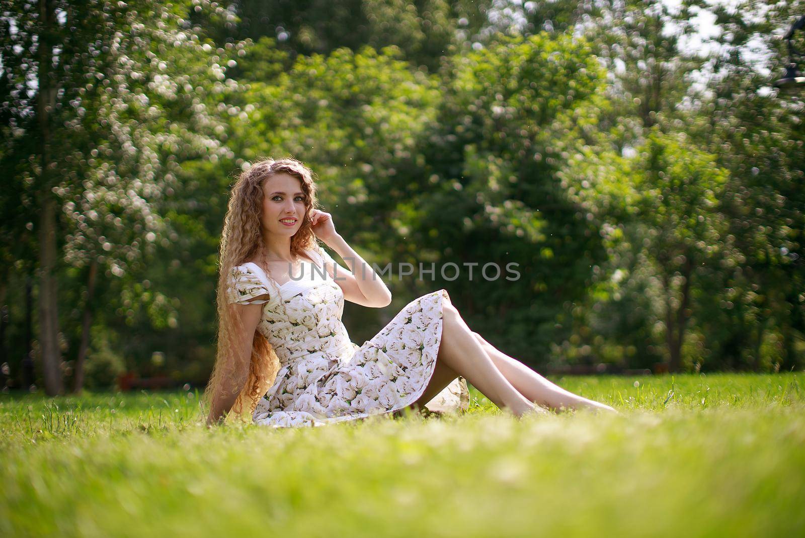Smiling blonde girl with long hair sitting on green grass. side view