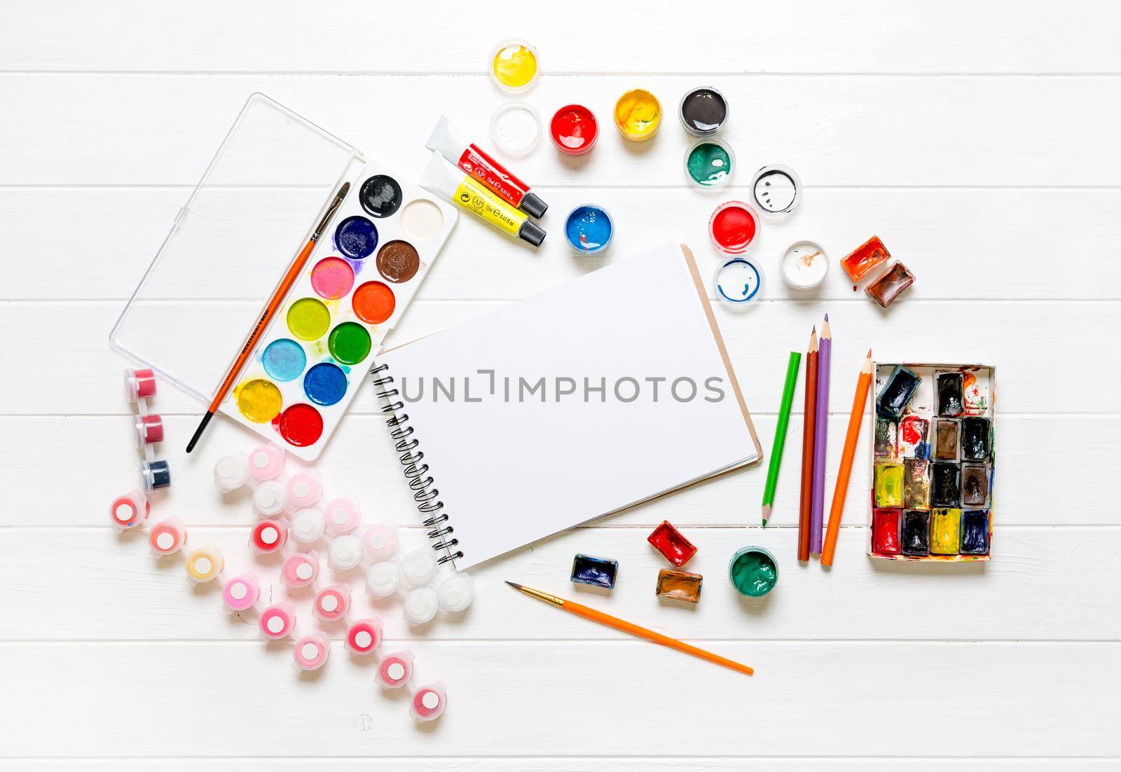White blank notepad among drawing accessories on white wooden background. Top view of color pencils, watercolors and brushes. Copy space