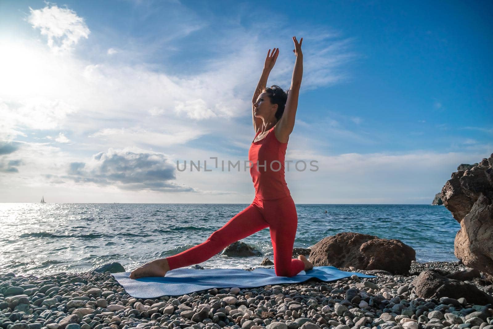 The woman in a red suit practicing yoga on stone at sunrise near the sea. Young beautiful girl in a red bathing suit sits on the seashore in lotus position. Yoga. Healthy lifestyle. Meditation