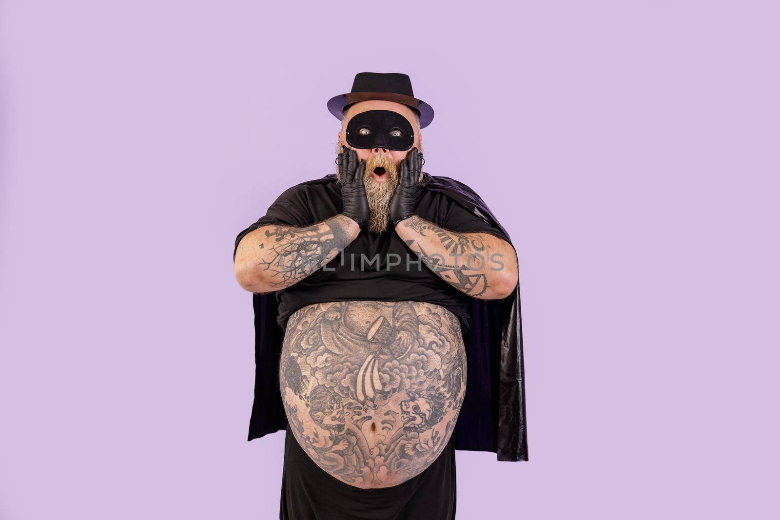 Shocked plump man in hero costume holds cheeks standing on purple background by Yaroslav_astakhov