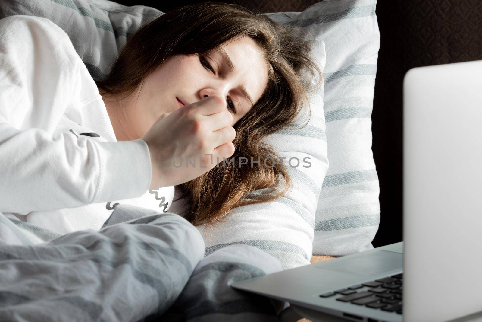 A young cute girl on a sunny day in a white jacket lies on the bed and looks at her nails. A laptop is standing nearby.