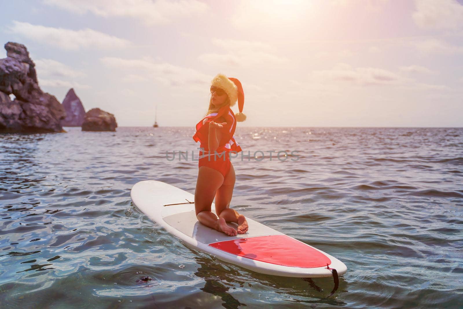 Young brunette woman in red swimsuit and Santa hat, swimming on kayak around basalt rocks like in Iceland. Back view. Christmas and travel concept