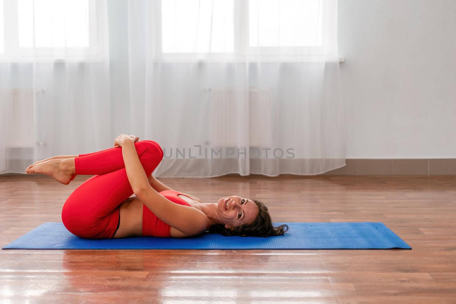 A young woman fitness instructor in red Sportswear Leggings and Top stretching in the gym before her pilates, on a yoga mat near the large window on a sunny day, female fitness yoga routine concept
